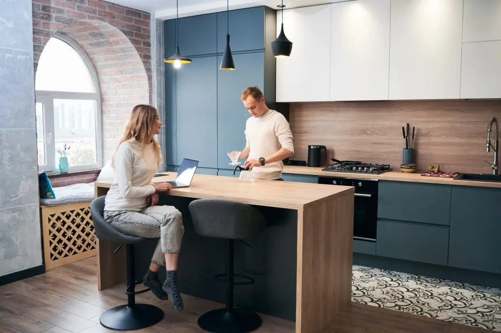 Side view of young woman and man working in laptop with hot tea in kitchen with beautiful modern furniture in blue color.
