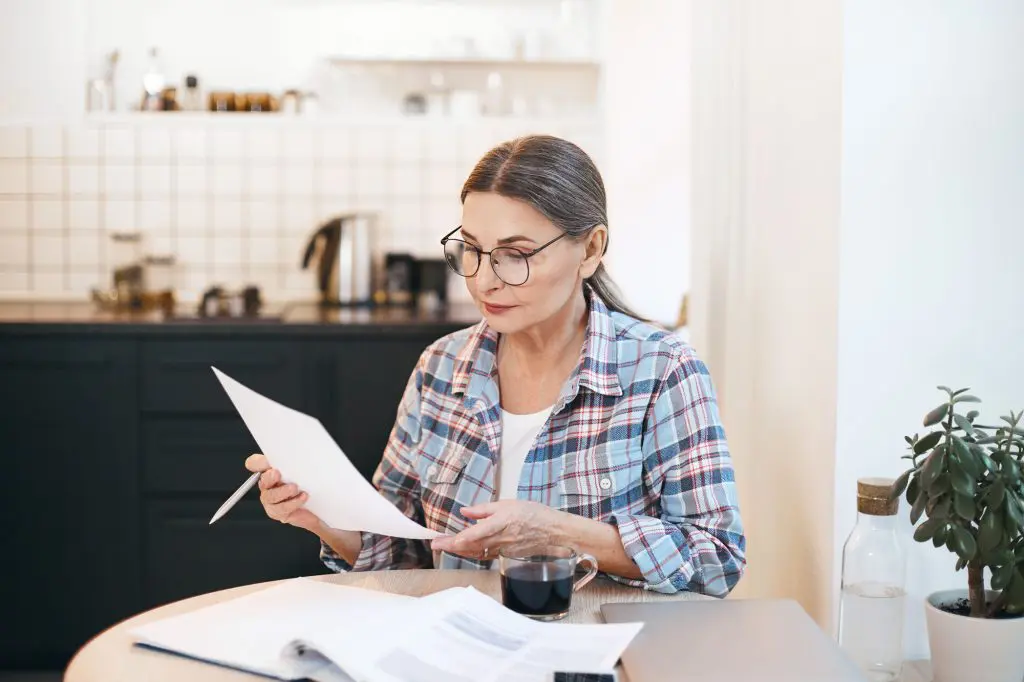 An older woman looks through papers as she reviews home remodel estimates at her kitchen table.
