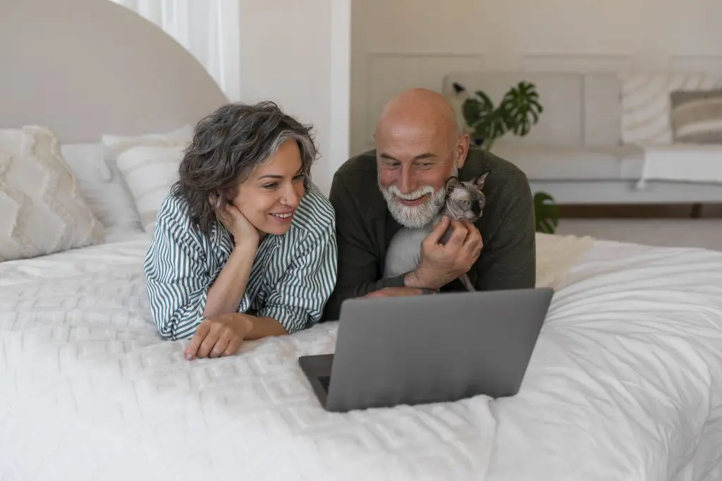A senior couple relaxes while watching a movie on their laptop in bed with their cat in their first-floor master suite.