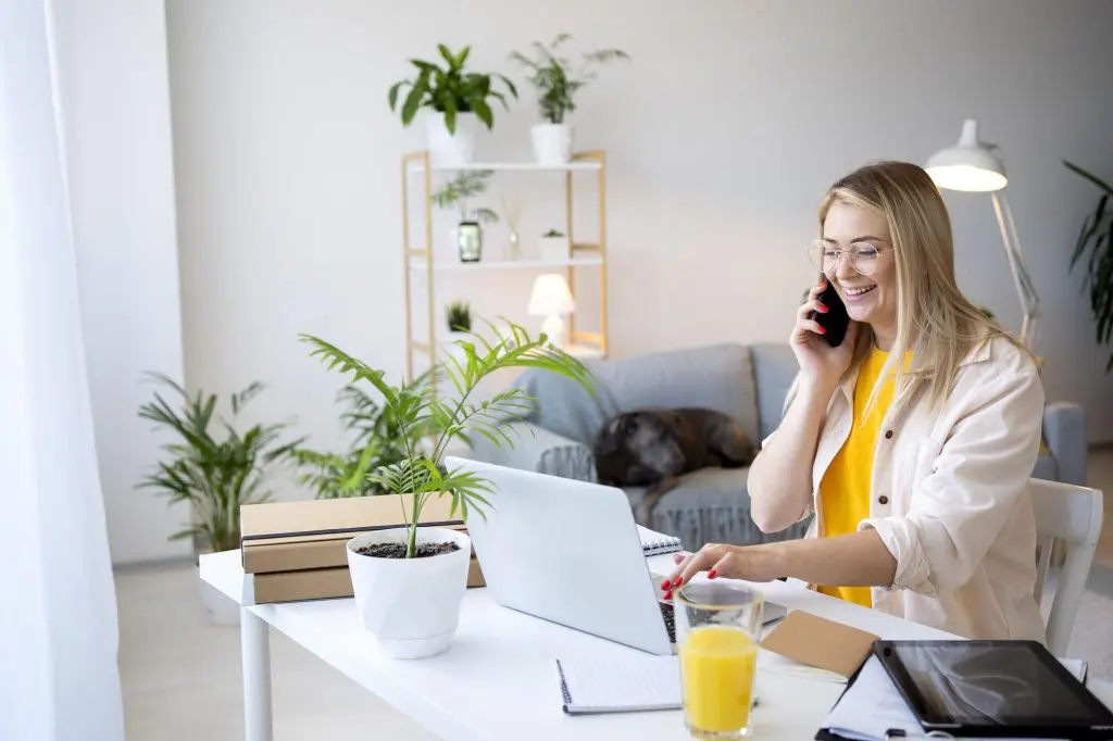 A woman smiles and talks on the phone while looking at her laptop. She sits at a desk in her modern, minimal home office.
