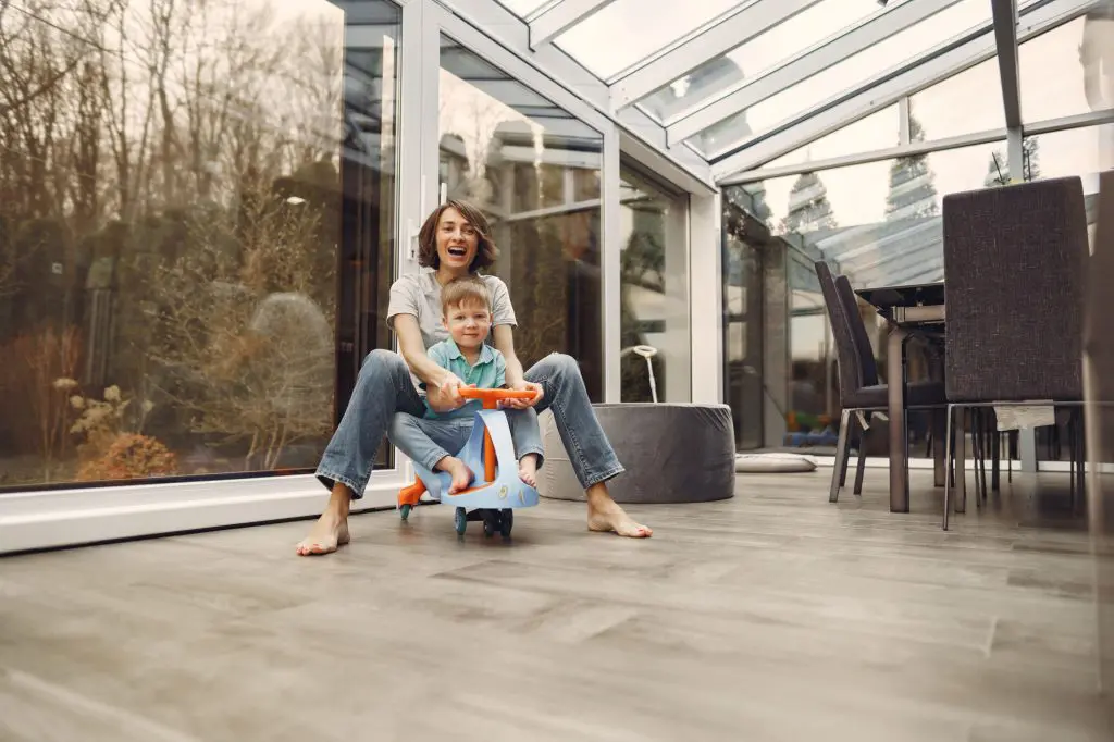 A woman in a gray t-shirt rides aroun on a toy car with her son in their homes newly added sunroom.