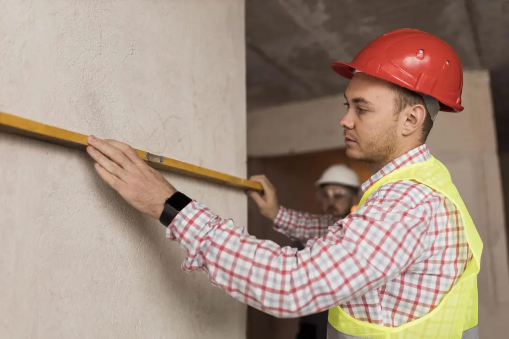 A contractor wearing a hardhat takes measurements of an interior wall during a home addition.